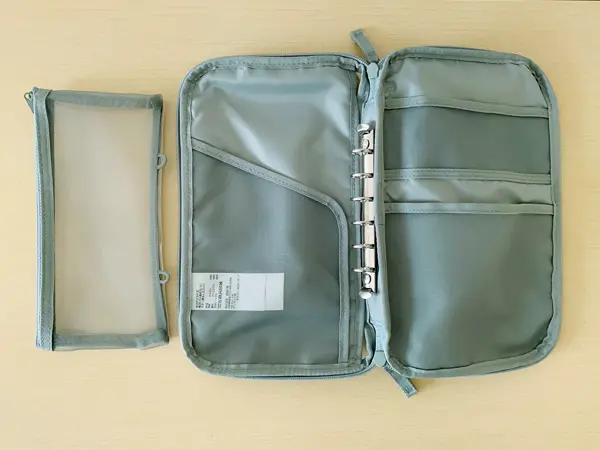 無印良品『コレクションケースA5スリム』の検証写真です。この商品の色は淡いグリーンで、ファスナーを開けて、中を展開した画像になります。中には3つのポケットと真ん中部分はバインダーになっており6つ穴のルーズリーフが挟めるようになっています。付属のメッシュ地の袋型のケースもバインダーに挟めるように工夫されています。