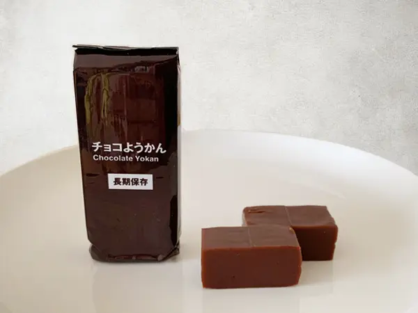 無印良品『備蓄おやつ チョコようかん』の検証写真です。1本単体のパッケージに入ったものと、実際に開けて切ったようかんがお皿に二つ並べてある画像です。ようかんの見た目は艶がなくチョコレートのお菓子みたいな印象です。